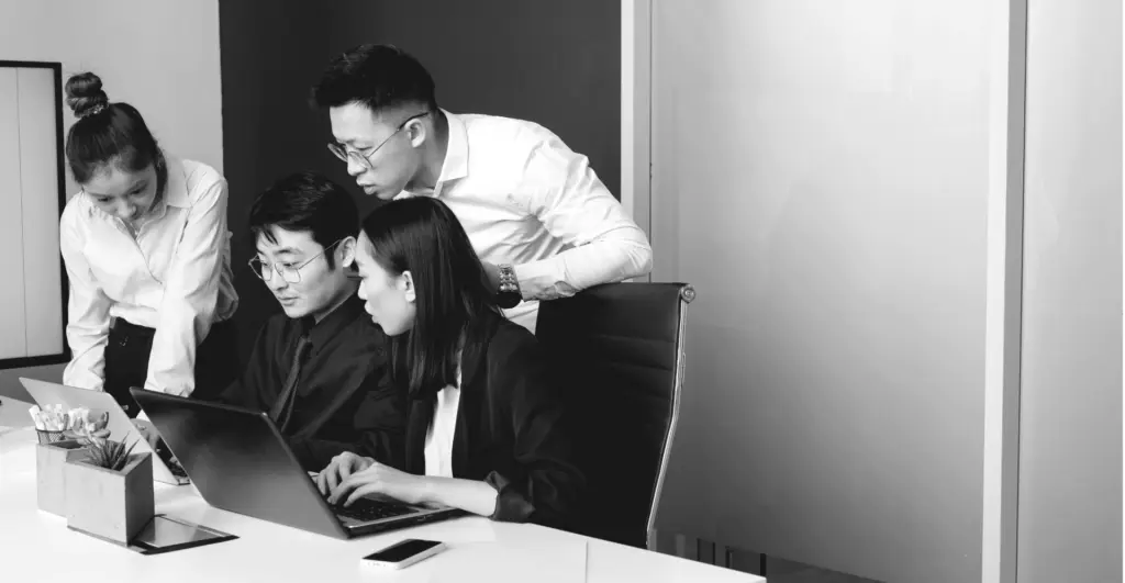 This image of corporate colleagues huddled around their work devices highlights the discussion topic: Ecommerce and Counterfeits in China with Sophia Lee.