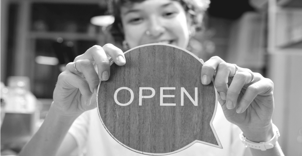 This image of a lady's happy face in front of the "open" sign for her business demonstrates our discussion topics: Facial recognition and the fight against impersonation scams.