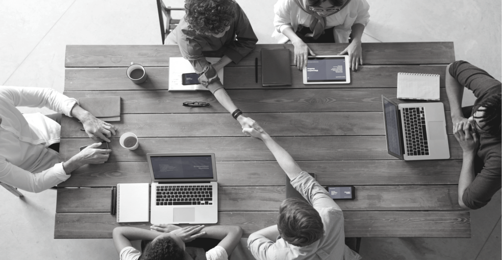 This image of colleagues shaking hands over a table where they word at their computers illustrates the conclusion of our overview of the pitfalls of ineffective brand protection, and how to correct them to protect your brand.